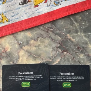 Presentkort  - Två presentkort från Systembolaget ligger på ett marmorbord. Ovanför syns en färgglad duk med tecknade figurer och en röd kant.