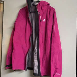 Rosa skaljacka från Peak Performance - Snygg rosa skaljacka från Peak Performance med justerbar huva och dragkedja framtill. Jackan har långa ärmar med kardborrejustering vid ärmsluten och är perfekt för utomhusaktiviteter. Märket 'Black Light' syns på ärmen.lite sliten i nacken på insidan men syns inte när man har på sig jackan. 