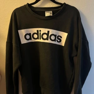 Svart sweatshirt från Adidas - Säljer en svart sweatshirt från Adidas med stort vitt logotryck på framsidan. Tröjan har långa ärmar och en rund halsringning. Perfekt för en avslappnad stil.