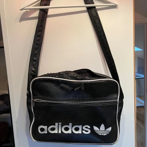 Svart axelväska från Adidas - Säljer en svart axelväska från Adidas med vit logga och detaljer. Väskan har en dragkedja och justerbar axelrem. Perfekt för en sportig look eller vardagsbruk.