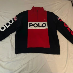 Tröja från Polo Ralph Lauren Sport - Snygg tröja från Polo Ralph Lauren i rött och mörkblå med stora vita och röda bokstäver 'POLO' på ärmarna och bröstet. Tröjan har en halv dragkedja och amerikanska flaggor på ärmarna. Perfekt för en sportig look!