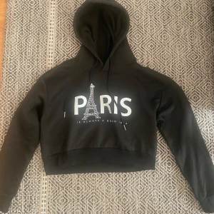 Svart croppad hoodie från Shein - Säljer en svart croppad hoodie från Shein med texten 'Paris' och Eiffeltornet tryckt på framsidan. Hoodien har långa ärmar och en bekväm huva. 