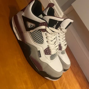 Nike Air Jordan 4 Paris Saint-Germain - Snygga Nike Air Jordans i samarbete med Paris Saint-Germain. Skorna har en stilren design i vitt, grått och vinrött med snörning och ikoniska detaljer. Perfekta för sneaker-entusiaster!