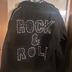 Svart jeansjacka med nitar - Cool svart jeansjacka med nitar som bildar texten 'Rock & Roll' på ryggen. Jackan har knappar framtill och nitar längs axlar och ärmar för en edgy look.