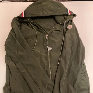 Grön vindjacka från Moncler - Snygg grön vindjacka från Moncler med huva och dragkedja. Jackan har Moncler-logga på armen och randiga detaljer i rött, vitt och blått på huvan. Perfekt för blåsiga dagar