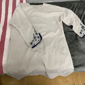 Vit abaya med broderade detaljer - Elegant vit abaya med vackra blå broderade blommor på ärmsluten. Klänningen har en enkel och stilren design med trekvartsärmar, perfekt för en avslappnad men chic look.