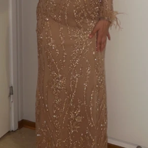 Glittrig beige långklänning - Elegant beige långklänning med glittriga detaljer och långa ärmar. Klänningen har en hög hals och ett bälte i midjan för en smickrande passform. Perfekt för festliga tillfällen.