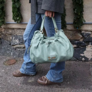 MaxMara Mintgrön handväska - Från MaxMara!! Snygg mintgrön handväska i syntetmaterial med praktisk dragkedja. Väskan har dubbla handtag och en dekorativ metallplatta framtill. Perfekt för att bära med sig det nödvändigaste.