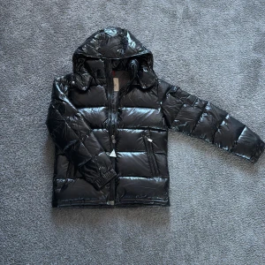 Svart glansig pufferjacka från Moncler - Snygg svart pufferjacka från Moncler med glansig finish och huva. Jackan har en dragkedja framtill och Moncler-logga på ärmen. Perfekt för kyliga dagar. Kom gärna med prisförslag, tags ingår ej