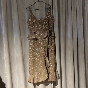 Brun prickig omlottklänning 38 - Charmig brun klänning med vita prickar från Make Way. Klänningen har en omlott-design med knytband i midjan och volangdetaljer. Perfekt för en somrig look!