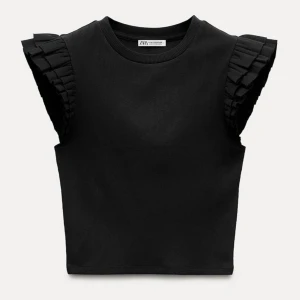 Svart ribbad topp från Zara - Snygg svart ribbad topp från Zara med coola volangdetaljer på axlarna. Perfekt för en stilren look, oavsett om du ska på fest eller bara vill känna dig extra fin till vardags. Toppen är ärmlös och har en tight passform som framhäver figuren.