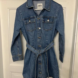 Jeansklänning från Zara - Säljer denna jeansklänning från Zara! Den är i en klassisk blå jeansfärg och har långa ärmar. Klänningen har knappar hela vägen ner och två bröstfickor med knappar. Den kommer med ett bälte i midjan för en snygg passform. Storlek S och inga defekter.