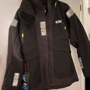 Svart jacka från Gill - Säljer en svart jacka från Gill. Den har en cool design med reflexdetaljer på ärmarna och bröstet. Jackan har en dragkedja framtill och är perfekt för blåsiga dagar. Den är långärmad och har en huva för extra skydd. 
