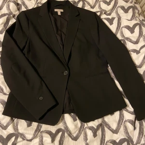 Kavaj - Helt ny svart kavaj från H&M med en knapp framtill och utan fickor. Kavajen har diskreta shoulder pads som ger en bättre form åt axlarna och en elegant, strukturerad siluett. Perfekt för en stilren och sofistikerad look!