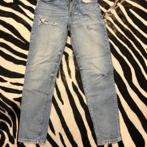 Blå jeans från Zara - Säljer ett par snygga blå jeans från Zara i storlek 30. De har en cool slitning på framsidan som ger dem en trendig look. Jeansen har en knappgylf och är i en klassisk straight fit. Perfekta för både vardag och fest!