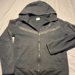 Svart hoodie från Nike - Säljer en svart hoodie från Nike med dragkedja och huva. Perfekt för en avslappnad stil. Den har långa ärmar och en diskret logga på bröstet.