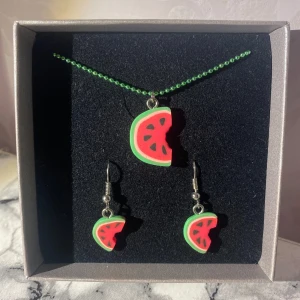 Halsband och örhängen med vattenmelonmotiv - Charmigt set med halsband och örhängen i form av vattenmelonskivor. Halsbandet har en grön kedja och både halsbandet och örhängena har färgglada detaljer i rött och grönt. Dom båda är oanvända!