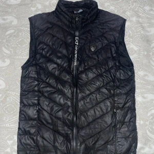 Svart väst från Emporio Armani - Snygg svart väst från Emporio Armani. Perfekt för lager-på-lager under kyliga dagar. Kommer med en praktisk förvaringspåse. Perfekt passform och väldigt stillig. Den är köpt på NK för 2000kr men jag har använt den ganska mycket så jag säljer den för 1000 priset är ej fast hugget i sten, hör gärna av dig om du har frågor. (Dålig bild, sök gärna upp den för att få se hur den ser ut bättre)