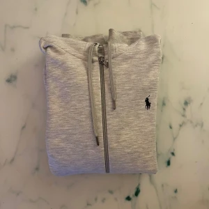 Grå Zip hoodie från Ralph Lauren - Snygg och bekväm grå hoodie från Ralph Lauren med dragkedja. Tröjan har en broderad logotyp på bröstet och praktiska fickor framtill. Perfekt för en avslappnad stil. 