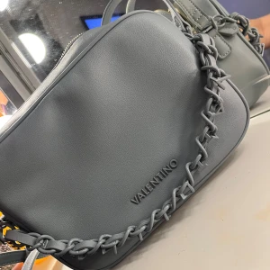 Grå axelväska från Valentino - Säljer nu min gråa oanvända axelväska från Valentino då jag aldrig fått den till användning. Den har ett unikt flätat axelband. Väskan har en dragkedja upptill och är tillverkad i skinn. 