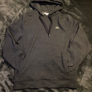 Mörkgrå hoodie från Lacoste - Säljer en stilren mörkgrå hoodie från Lacoste med klassisk passform. Tröjan har en justerbar huva med dragsko och det ikoniska Lacoste-logotypen på bröstet. Perfekt för en avslappnad look.