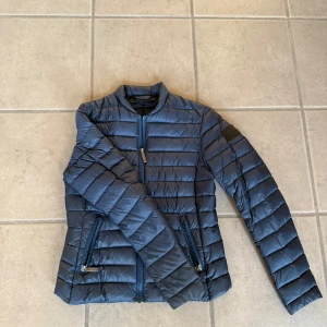 Blå dunjacka från Peak Performance - Snygg blå dunjacka från Peak Performance med dragkedja och quiltad design. Perfekt för kyligare dagar med sin lätta och värmande konstruktion. Jackan har en stilren look med diskret logga på ärmen.