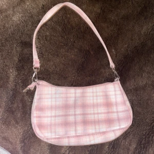 Rutig gammel rosa axelväska - Söt rosa axelväska med rutigt mönster och dragkedja. Perfekt för att bära med sig det viktigaste. Väskan har en enkel design med en justerbar axelrem. Rätt så liten, Har något som jag tror är en vattenfläck men borde gå bort i tvärtt. Väldigt omärkbar fläck.