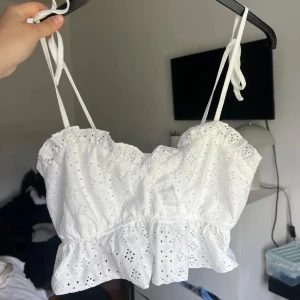 Vit broderad topp med volang - Charmig vit topp med broderade detaljer och volangkant. Axelbandslös design med knytband för justerbar passform. Perfekt för sommarens varma dagar.