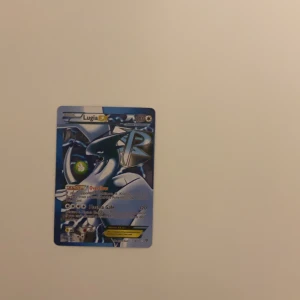 Lugia EX Pokémonkort - Säljer ett Lugia EX Pokémonkort med en snygg design och holografiska detaljer. Perfekt för samlare eller spelare som vill stärka sin kortlek. Kortet har en blå och silver färgpalett med Lugia i en kraftfull pose.