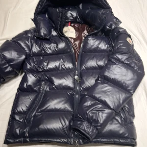  Moncler Maya - Säljer en snygg och varm mörkblå dunjacka från Moncler. Jackan har en dragkedja framtill och en huva för extra skydd mot kyla. Insidan är brun och har ett unikt mönster. Perfekt för kalla dagar!