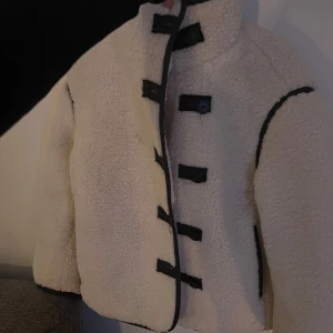 Populär teddyjacka hm  - Mysig teddyjacka från H&M med svarta knappar och detaljer. Jackan har en hög krage och långa ärmar. Den är stilren och bekväm!