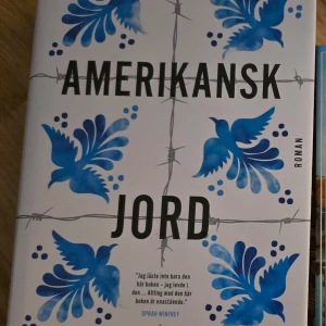 Amerikansk jord - Romanen 'Amerikansk jord' har ett omslag med blå blommönster och taggtrådsmotiv. Citat från Oprah Winfrey finns på framsidan.