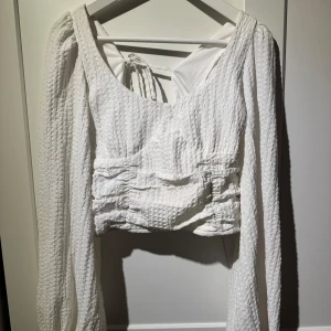 Vit blus med knytning - Säljer en stilren vit blus med långärmad design och knytning i ryggen. Blusen har en texturerad yta och en v-ringad framdel som ger en elegant touch. Perfekt för både vardag och speciella tillfällen.