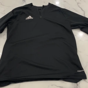 Svart långärmad tröja från Adidas - Säljer en svart långärmad tröja från Adidas med dragkedja vid halsen. Tröjan är tillverkad i Aeroready-material som andas, perfekt för träning. Adidas-loggan är tryckt på bröstet.