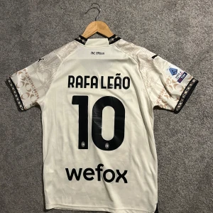 AC Milan fotbollströja med Rafa Leão 10 - Snygg AC Milan pique fotbollströja med Rafa Leão nummer 10. Tröjan är vit med ett unikt mönster och har korta ärmar. Perfekt för fans av laget och spelaren!