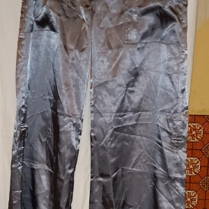 Grå byxor i polyester - Snygga grå byxor i glansigt polyester med en loose passform. Byxorna har dekorativa ringdetaljer på sidorna och är perfekta för en stilren look. Storlek 42. Går att justera benlängden. Dessa kommer också från E18shoppen