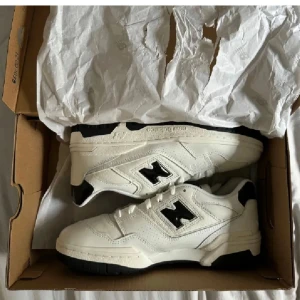 Vita sneakers från New Balance - Snygga vita sneakers från New Balance med svart detalj på sidan. Skorna har en klassisk design med snörning och en bekväm sula. Perfekta för en stilren look.