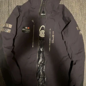  Svart Sail Racing jacka - Snygg svart jacka från Sail Racing med dragkedja och huva. Jackan har flera logotyper och detaljer på bröstet och ärmarna. Perfekt för kyliga dagar.