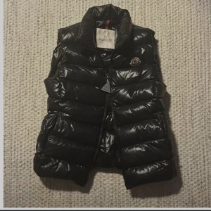 Svart dunväst från Moncler - Snygg svart dunväst från Moncler med hög krage och dragkedja. Perfekt för kyliga dagar när du vill hålla stilen. Västen har en glansig finish och Moncler-logga på bröstet.