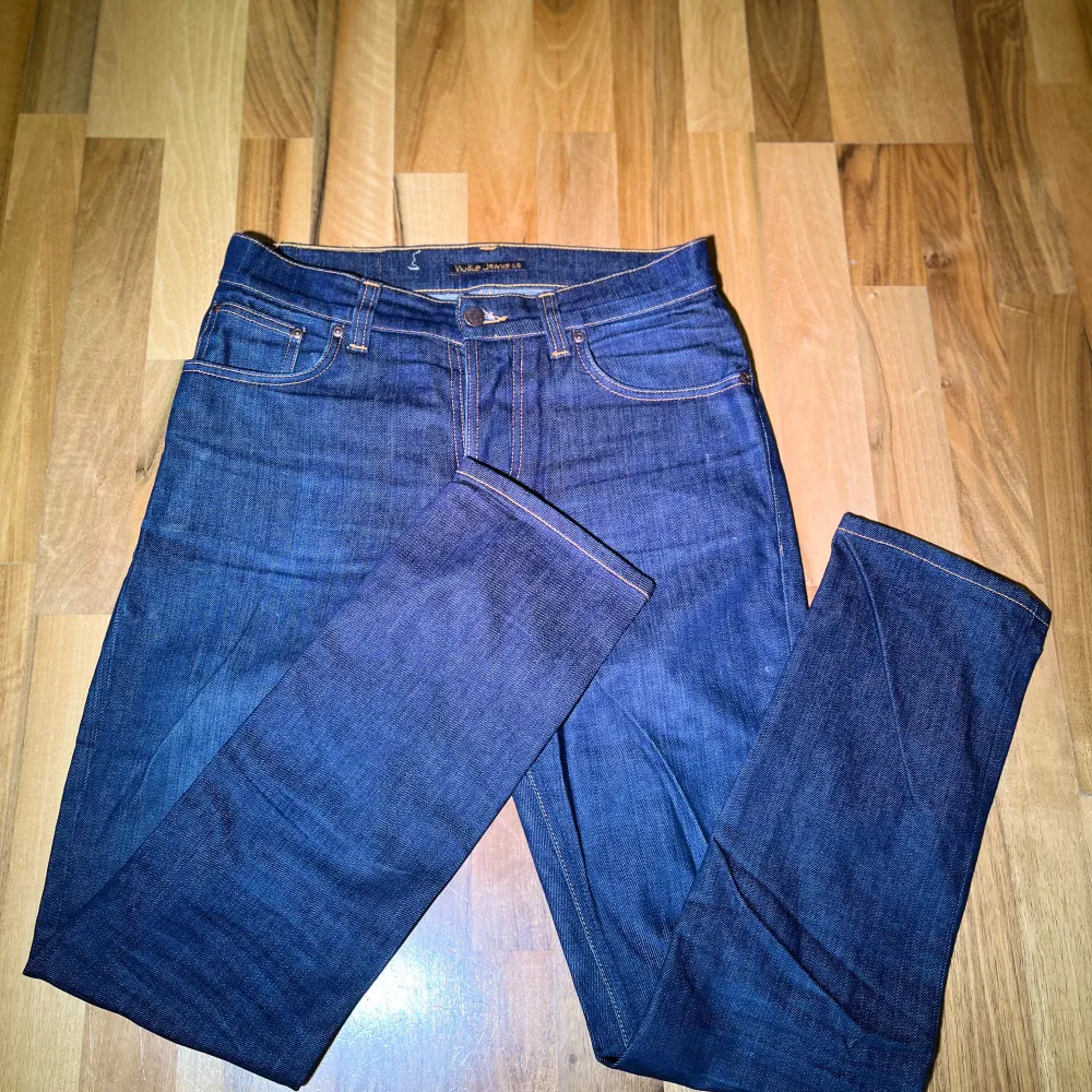 Säljer ett par mörkblå jeans från Nudie Jeans med klassisk femficksdesign och orange sömmar. Perfekta för en stilren look. Model: grim tim, storlek 31/32, finns små fläckar som ej syns på en distans. Farkut & Housut.