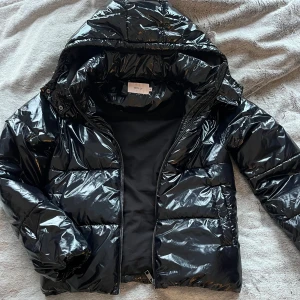 Svart glansig pufferjacka från Nelly - Snygg svart pufferjacka från Nelly med glansig finish och huva som är helt slutsåld. Jackan har en dragkedja framtill och är perfekt för kyligare dagar. Den är både stilren, praktisk och sparsamt använd🤍som ny!