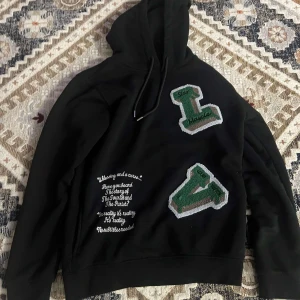 Svart hoodie från Louis Vuitton - Snygg svart hoodie från Louis Vuitton med gröna och vita bokstäver på framsidan. Den har en klassisk huva med dragsko och textdetaljer i vitt. Perfekt för en stilren och avslappnad look.
