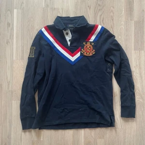 Mörkblå pikétröja från Polo Ralph Lauren - Snygg mörkblå långärmad pikétröja från Polo Ralph Lauren med färgglada ränder i rött, vitt och blått över bröstet. Tröjan har en broderad emblem på vänster sida och knappar vid kragen. Tröjan är i väldigt bra skick.