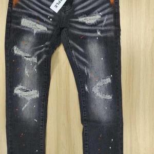 Svarta jeans med slitningar och färgstänk - Coola svarta jeans från Purple med slitningar och unika färgstänk i orange och vitt. Jeansen har en straight passform och är perfekta för en avslappnad stil. De har klassiska fem fickor och knappgylf.