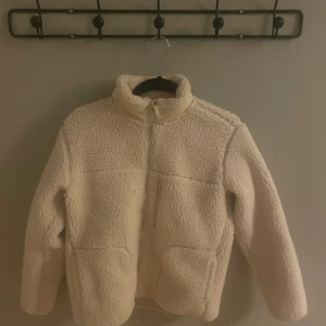 Vit teddyjacka från Uniqlo - Mysig vit teddyjacka från Uniqlo för barn. Jackan har en dragkedja framtill och är tillverkad i mjuk polyester. Perfekt för kyliga dagar med sin fluffiga och varma design.