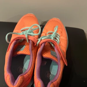 Orange sneakers med turkosa detaljer - Snygga och färgglada sneakers i orange med turkosa snören och detaljer. Skorna har en bekväm passform och är perfekta för en aktiv livsstil. De har även en praktisk kardborreknäppning för enkel på- och avtagning.