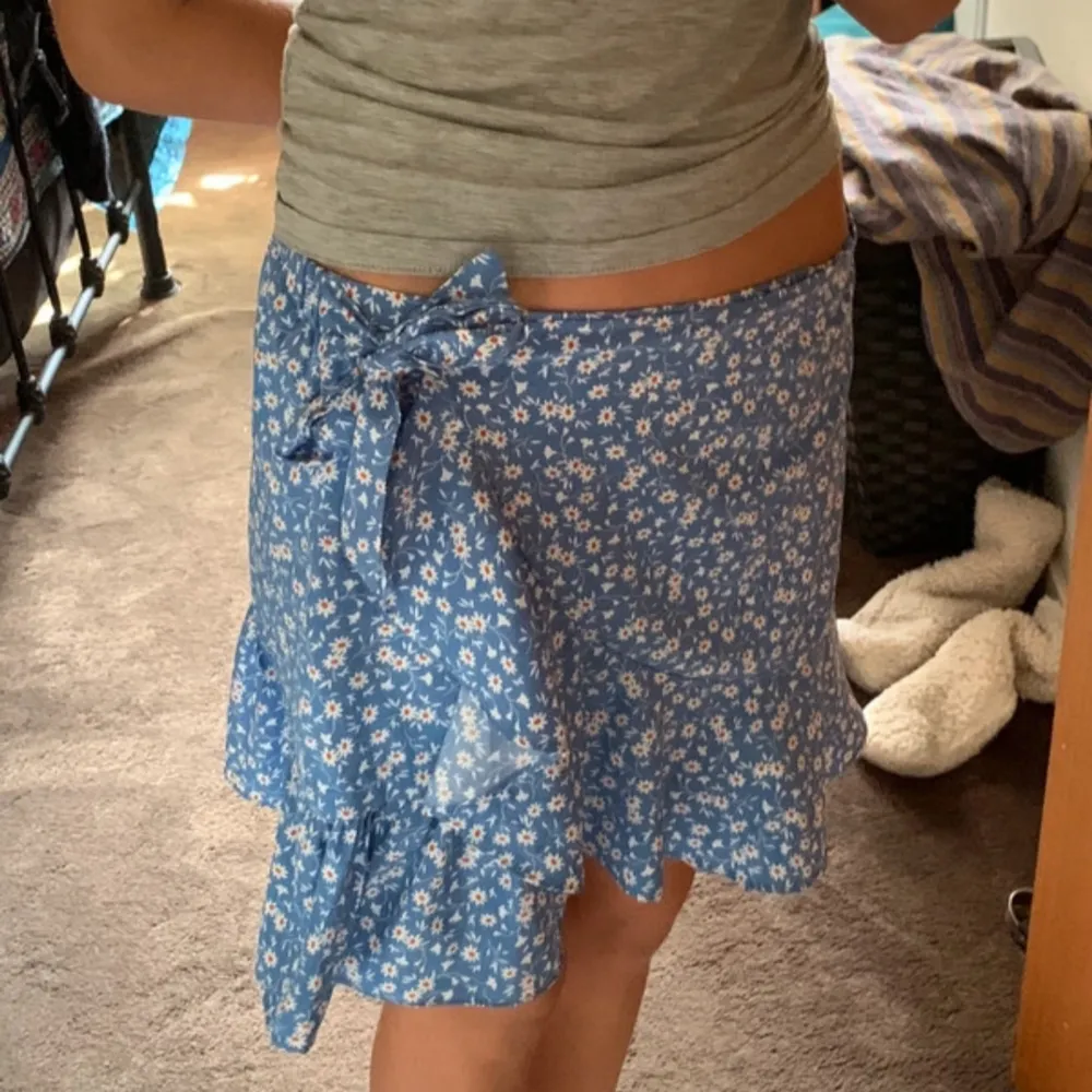 Söt blå omlottkjol med vita blommor och volangdetaljer. Kjolen har en knytning i midjan och är perfekt för varma dagar.. Hameet.