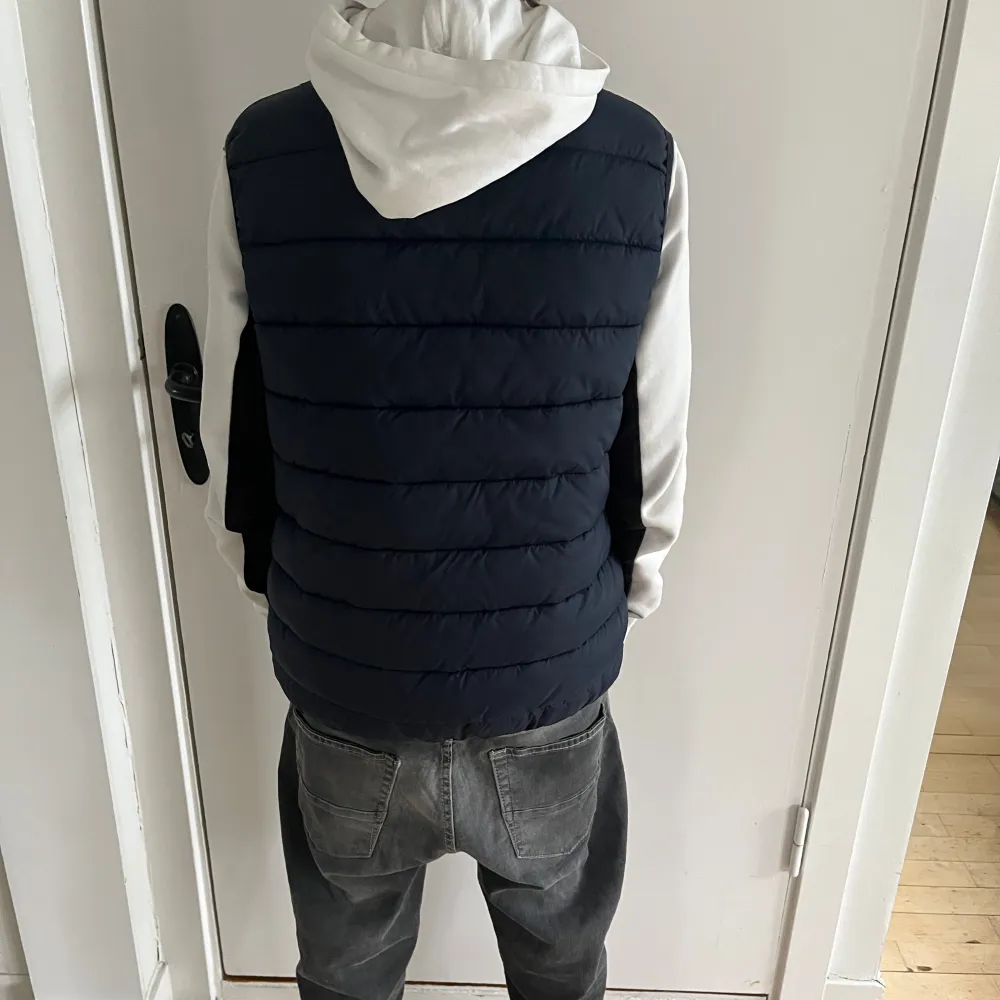 Säljer en snygg mörkblå barn dunväst med dragkedja framtill. Västen har en liten logga på bröstet och är perfekt för kyliga dagar. Den är både stilren och praktisk, idealisk för höst och vår.. Takit.