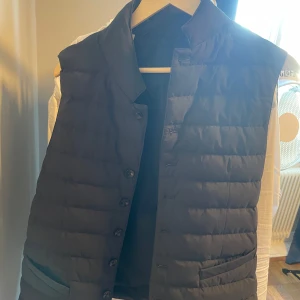 Suitsupply väst marinblå - Snygg marinblå dunväst från Suitsupply med knappar framtill. Västen har en stilren design med två fickor och en hög krage. Perfekt för lager-på-lager under kyligare dagar eller något utanför en tröja på varmare dagar.