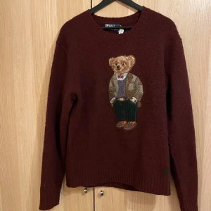Vinröd tröja med nallemotiv från Polo Ralph Lauren - Säljer en vinröd tröja från Polo Ralph Lauren med ett broderat nallemotiv på framsidan. Tröjan har långa ärmar och en rund halsringning. Perfekt för en mysig och stilren look.